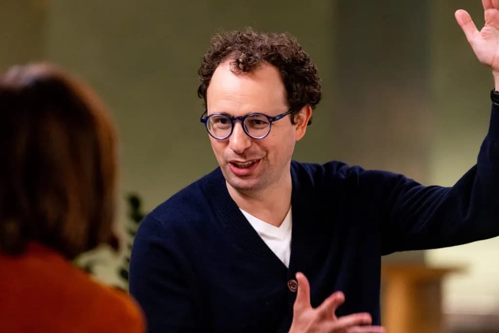 Anthropic CEO Dario Amodei gesturing with his hand raised while speaking during an interview, wearing round glasses and a navy cardigan