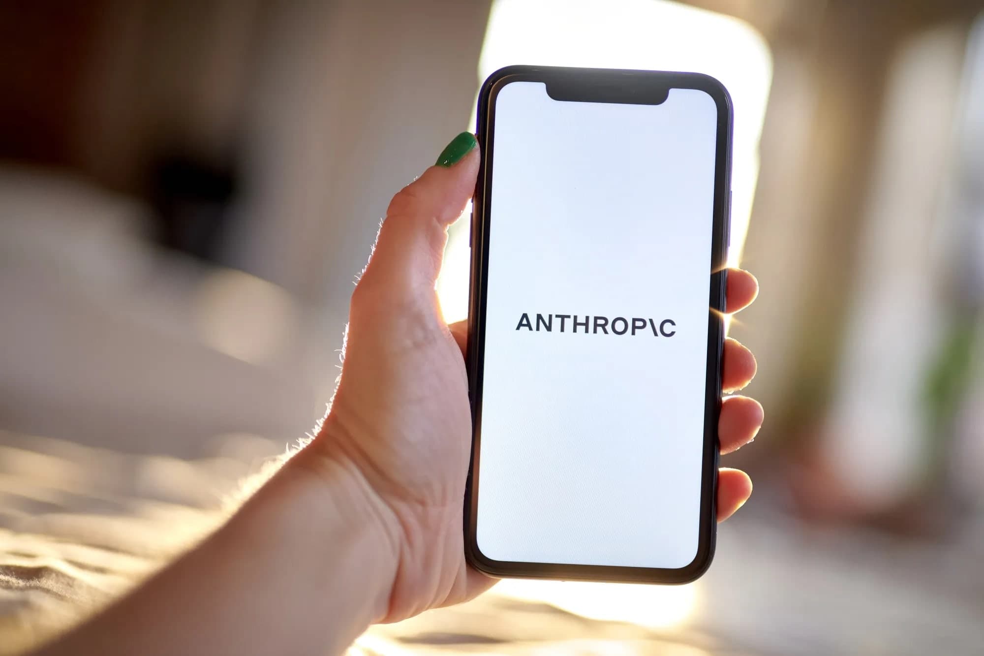 A hand holding a smartphone displaying the Anthropic logo on a white screen, with warm sunlit light in the background