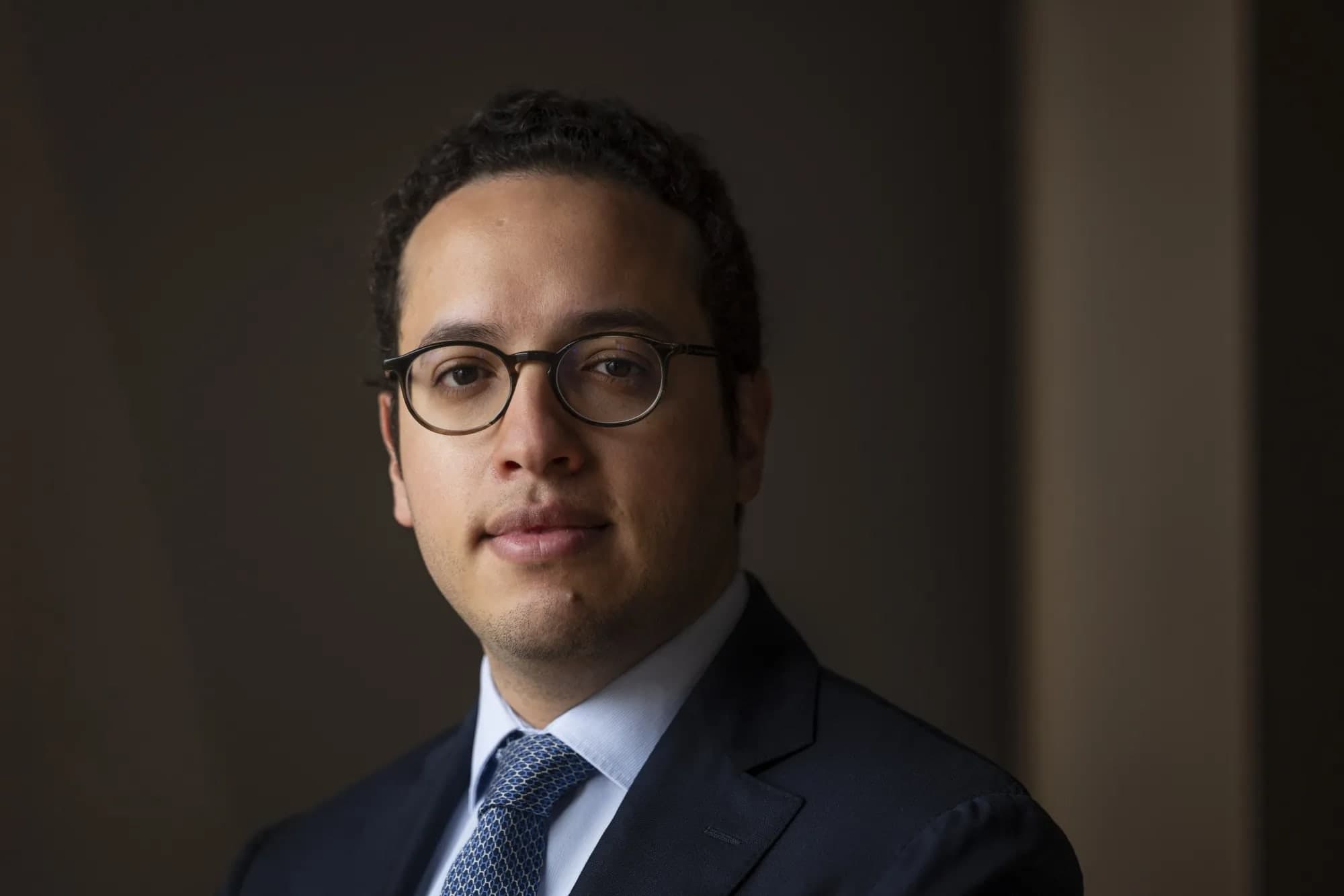A portrait of a man in glasses and a dark suit with a patterned blue tie, photographed against a dark brown backdrop