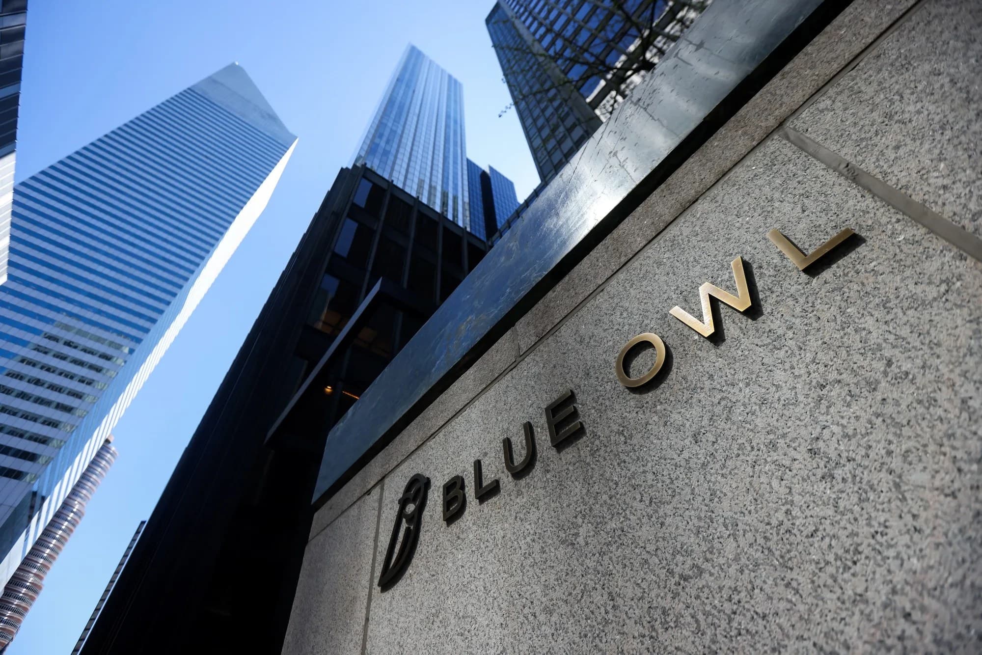 Low angle view of the Blue Owl sign carved into a stone building facade in New York City, with skyscrapers rising behind it against a blue sky