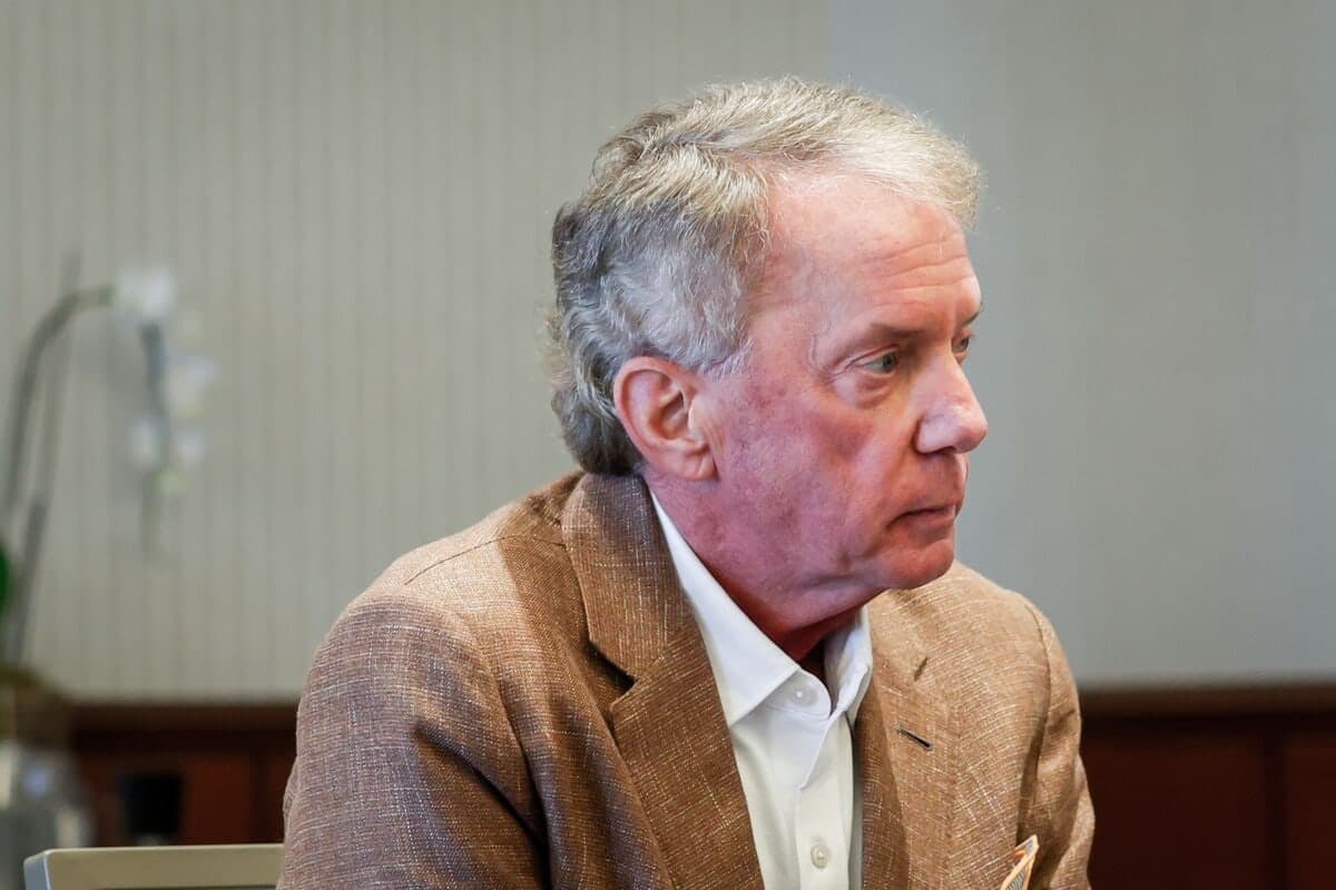 A man in a tan blazer sitting and looking to the side during an interview, with a wallpapered wall and a plant blurred in the background