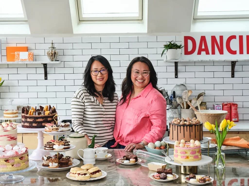 Sisters Annabel and Emily Lui standing together in the Cutter and Squidge kitchen, surrounded by tiered cakes, brownies, scones, and pastries on display stands