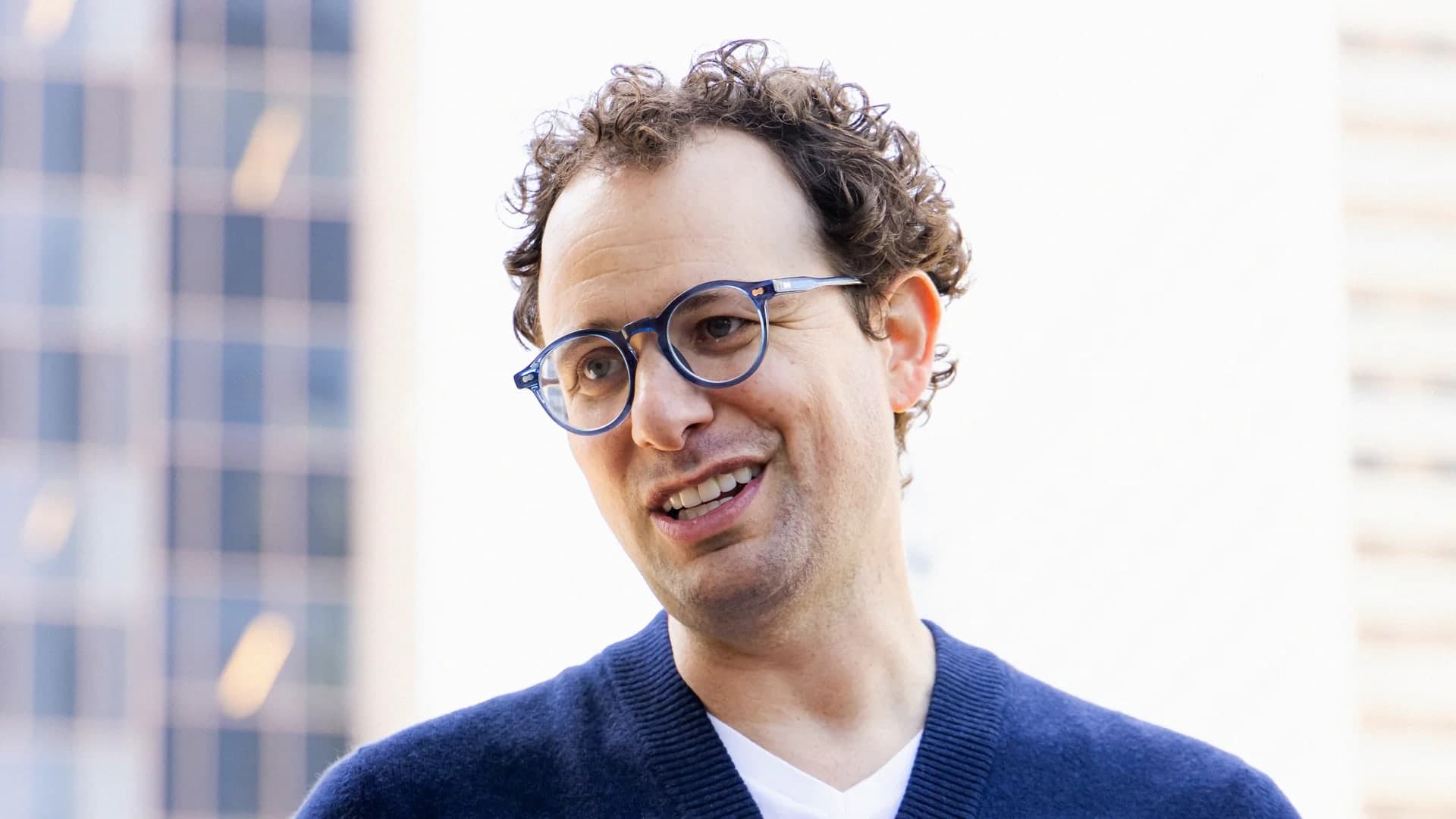 Dario Amodei smiling and speaking outdoors, wearing round glasses and a navy cardigan over a white shirt, with office buildings blurred in the background
