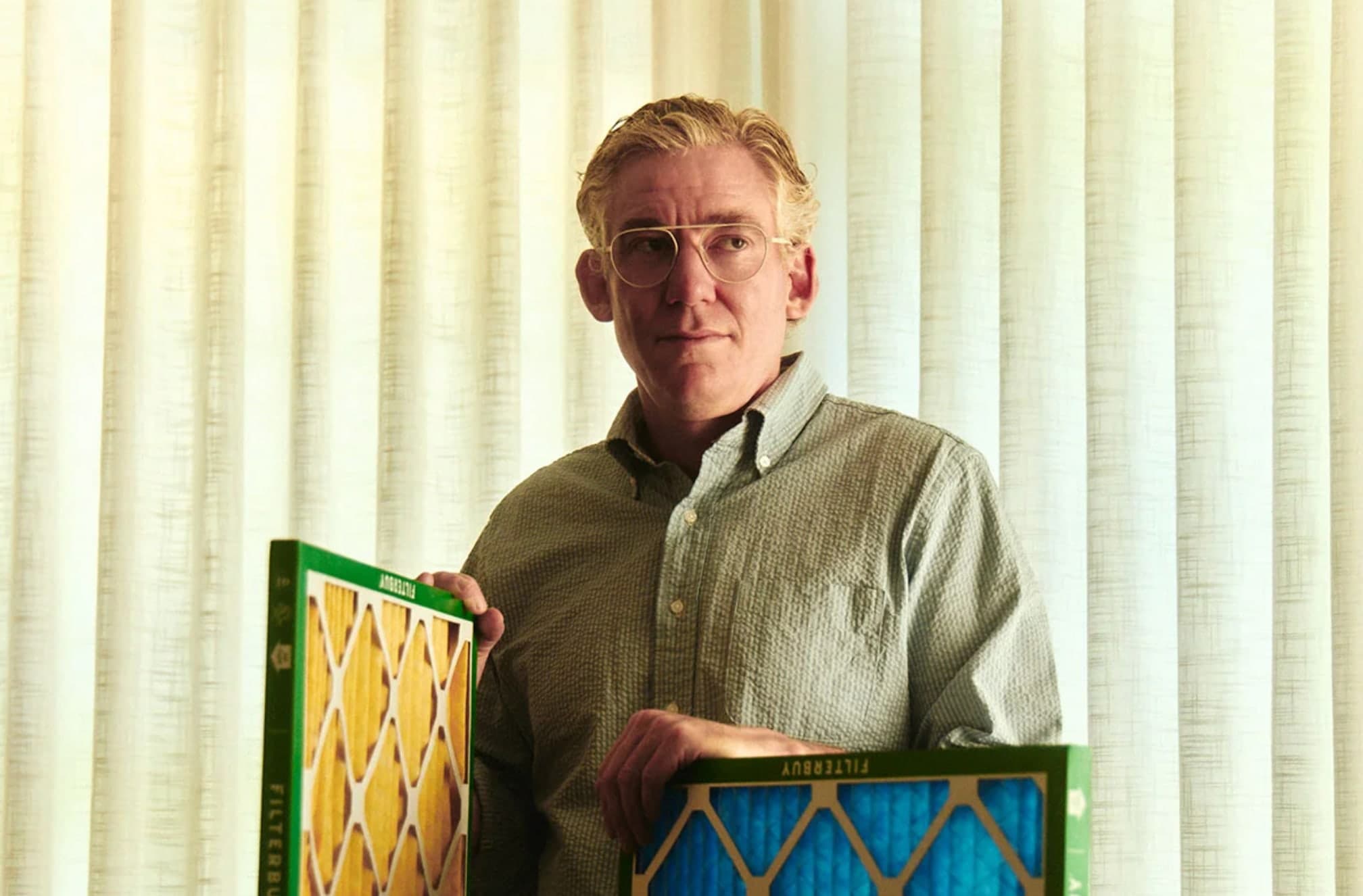 David Heacock standing in front of a curtain, holding two Filterbuy branded air filters, wearing round glasses and a green button up shirt