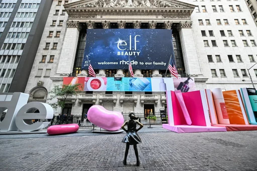 A large e.l.f. Beauty banner welcoming Rhode to the family displayed on the facade of the New York Stock Exchange, with oversized pink sculptures of beauty products on the street below