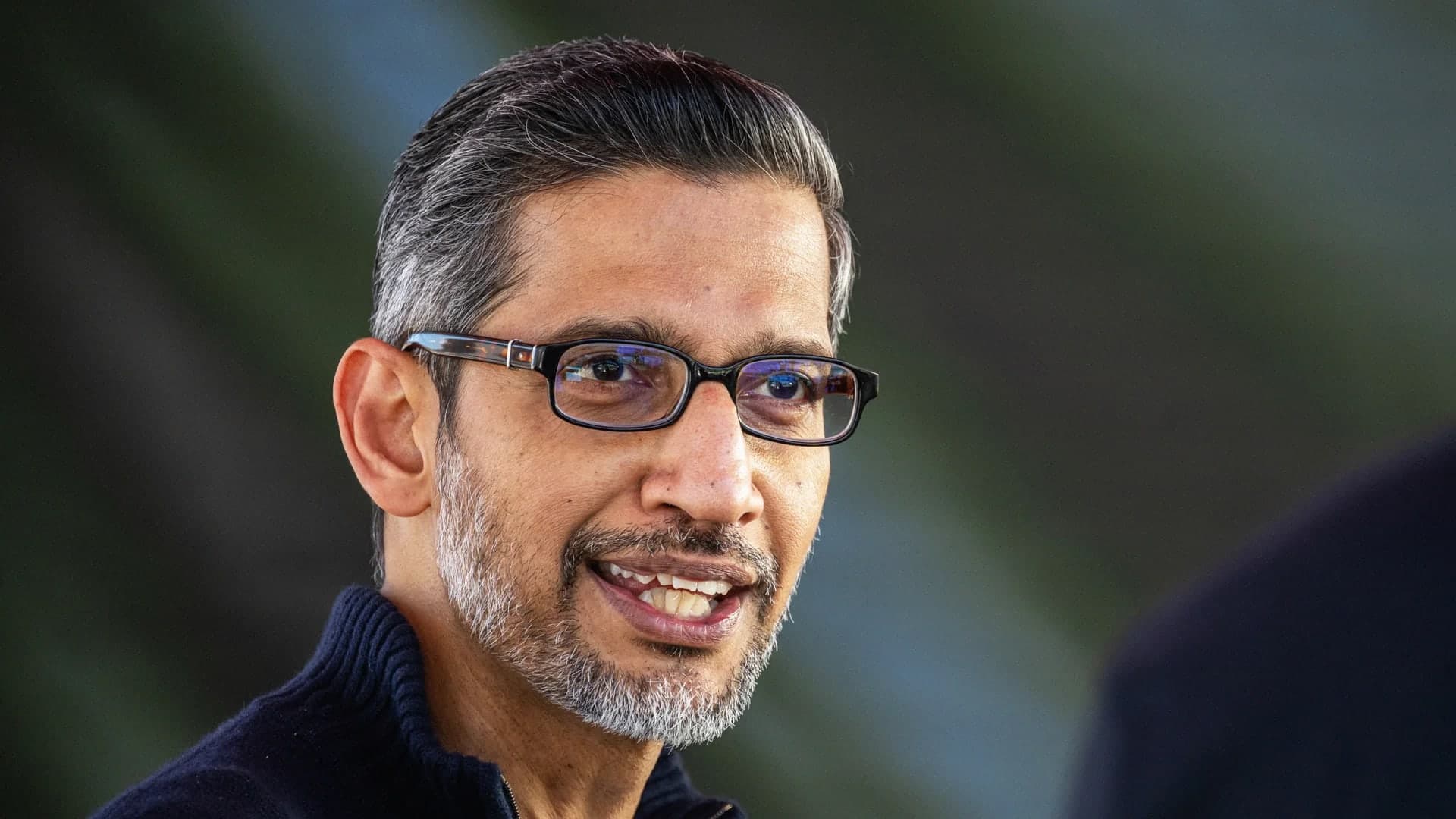 Sundar Pichai smiling outdoors, wearing rectangular glasses and a dark quarter zip sweater, with green foliage blurred in the background