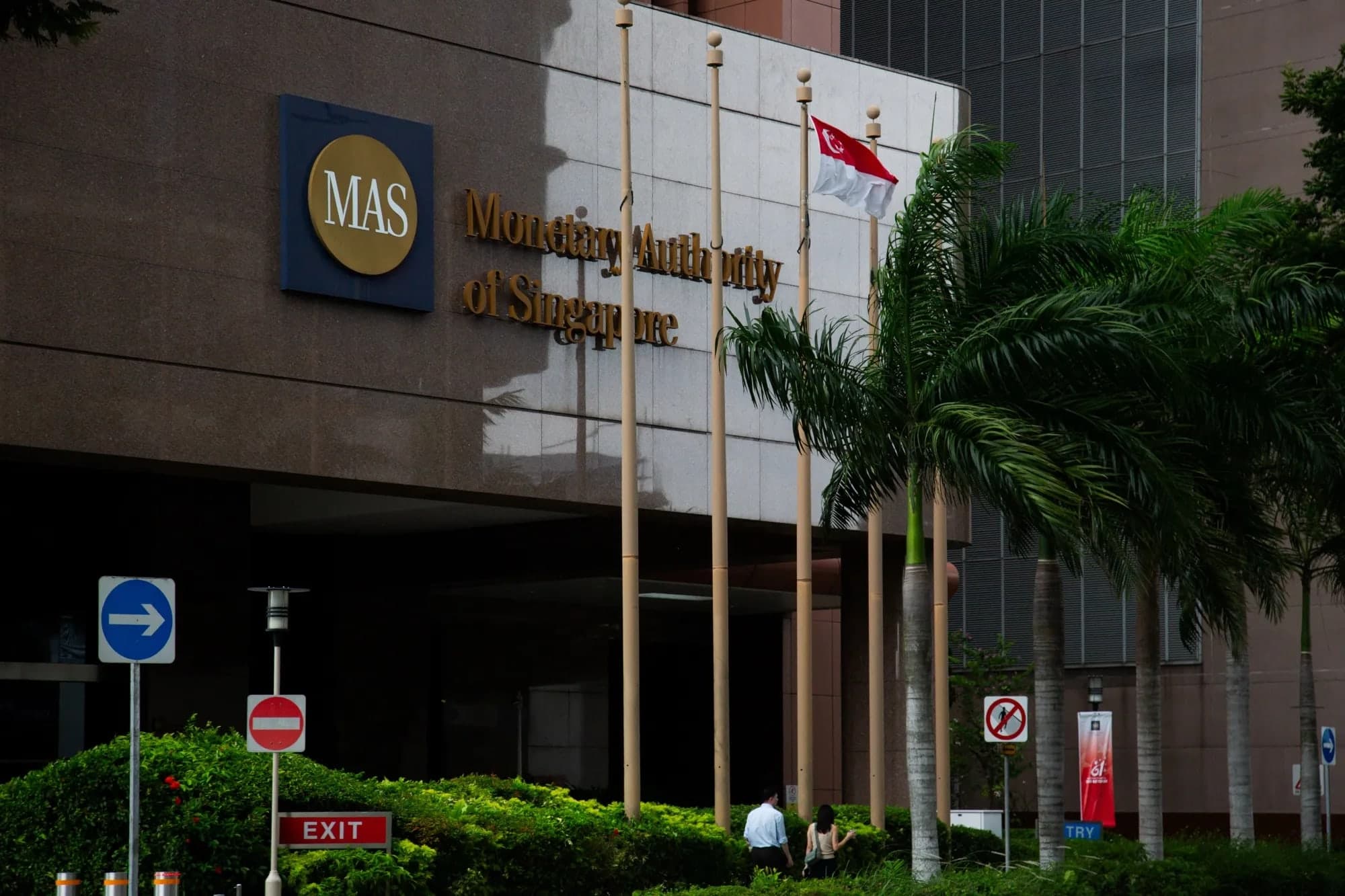 The Monetary Authority of Singapore sign and entrance on the facade of its headquarters building, with palm trees and Singapore flags in front