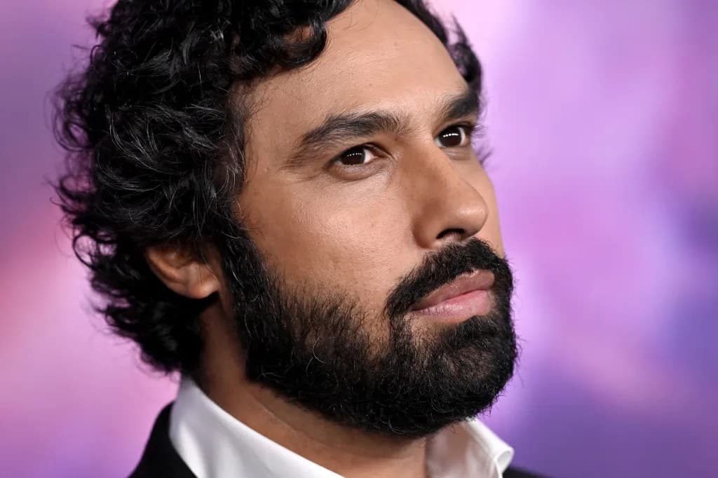 Close-up portrait of actor Kunal Nayyar with curly dark hair and a full beard, wearing a black suit and white shirt against a purple backdrop