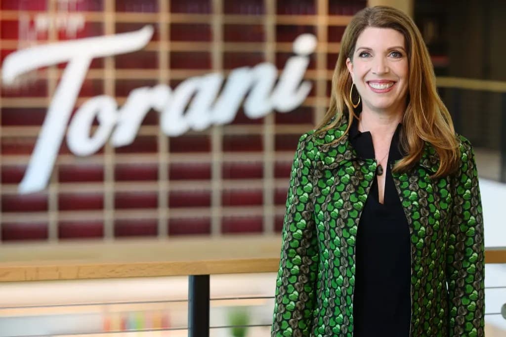 Melanie Dulbecco, wearing a green patterned jacket, smiling in front of the Torani logo at the company's headquarters