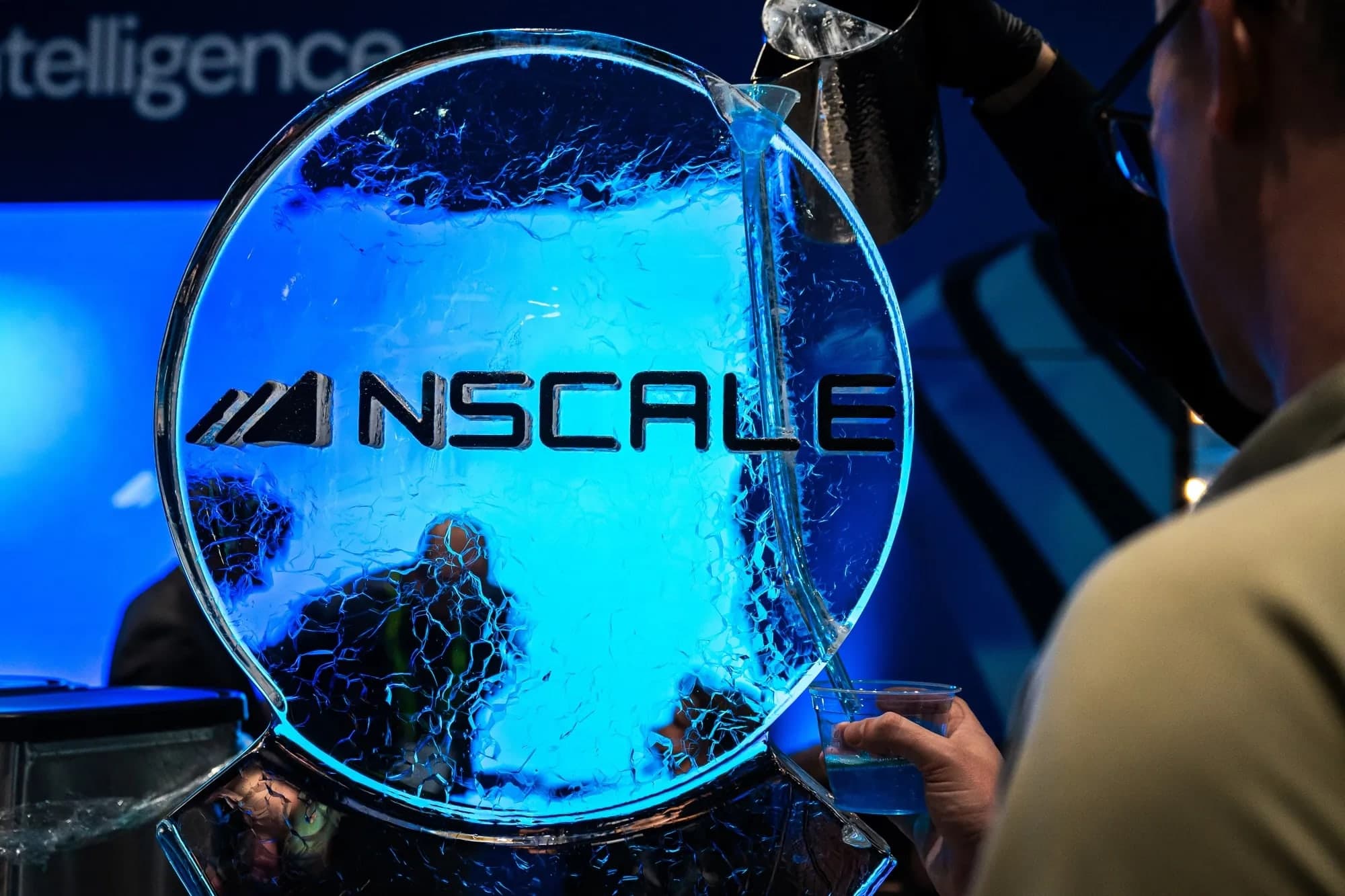 An illuminated blue ice sculpture engraved with the Nscale logo at an event, with a person holding a drink beside it