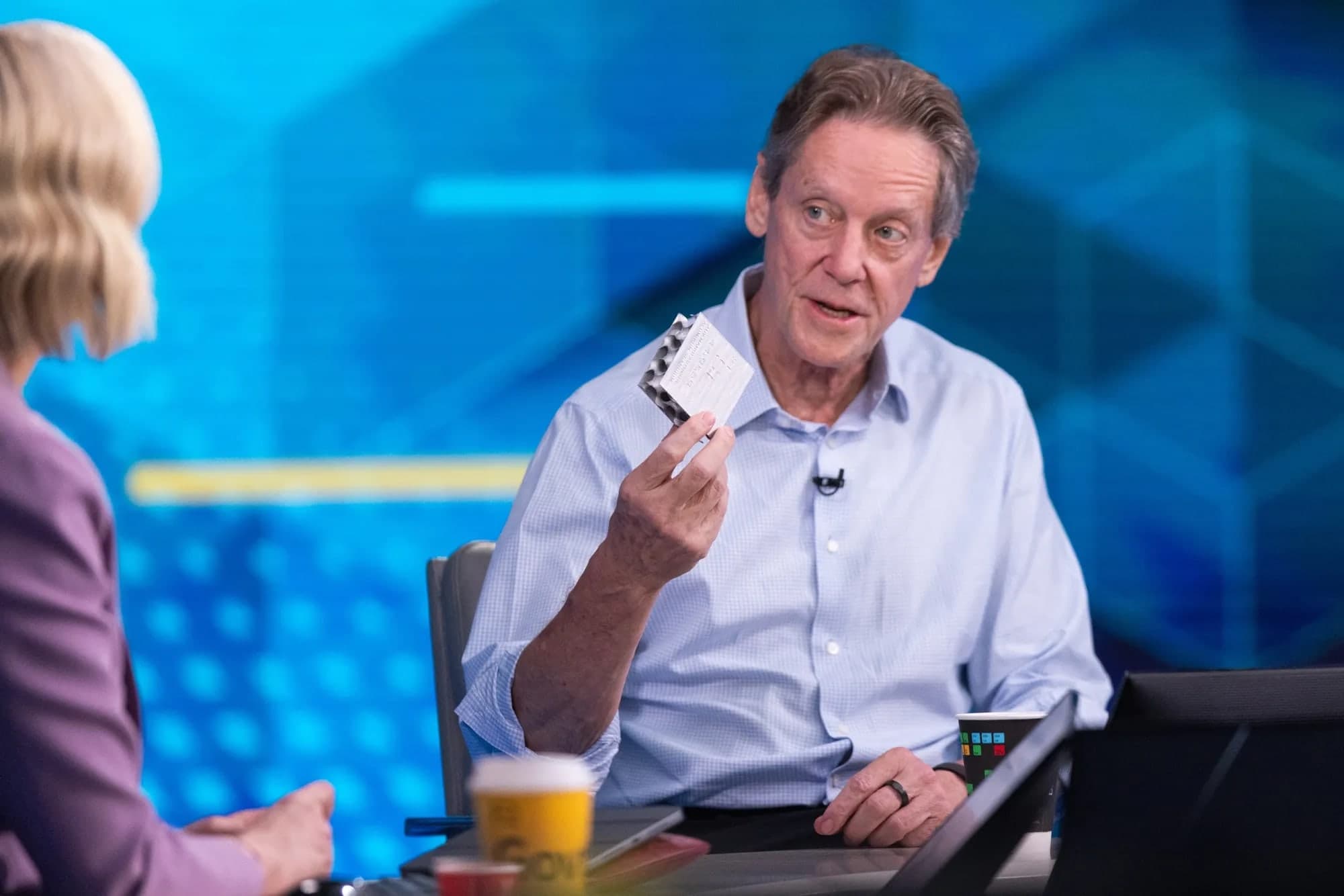 Robert Friedland holding a piece of paper while speaking during a television interview, seated at a desk with a coffee cup and laptop