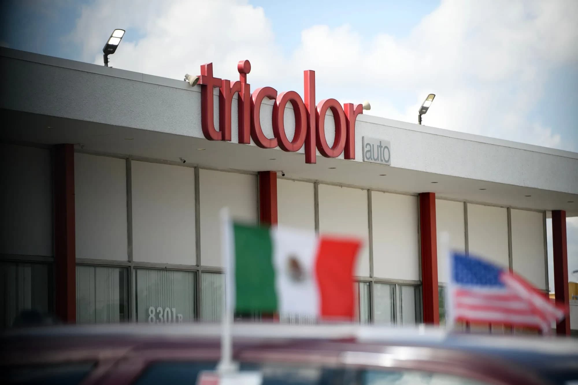 The Tricolor Auto dealership sign on a building facade, with Mexican and American flags visible in the foreground