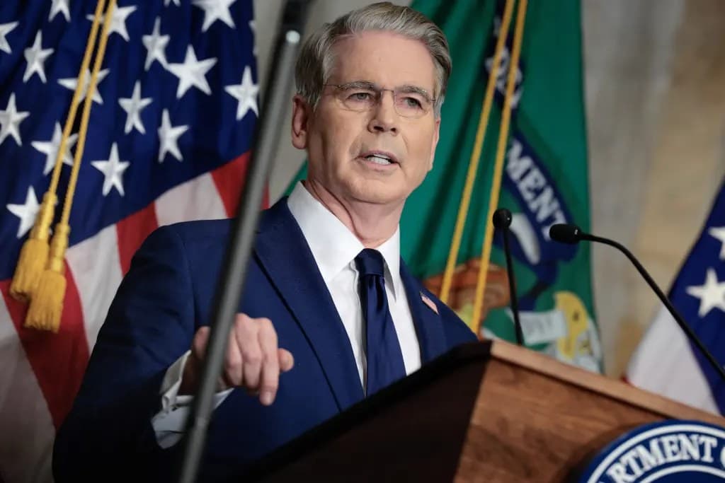 A man in a dark blue suit and glasses speaking and pointing while standing at a Department of the Treasury podium with American and Treasury flags behind him