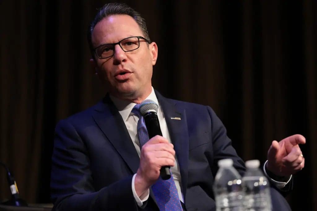 A man in a dark suit and glasses speaks into a microphone while gesturing with his other hand, seated at a table with two water bottles in front of him