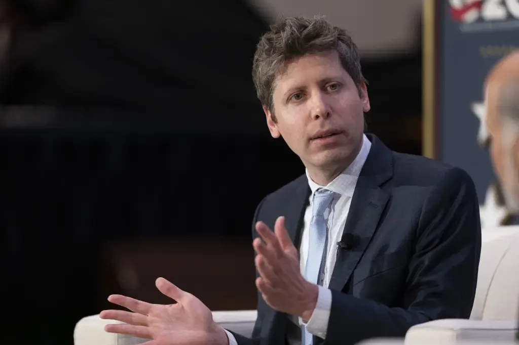 Sam Altman gestures with both hands while speaking on stage at a conference, wearing a dark suit and light blue tie