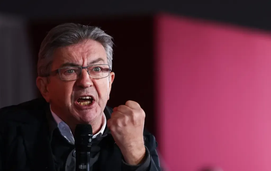 Jean-Luc Melenchon speaks forcefully into a microphone with a raised, clenched fist, set against a dark background with a pink accent