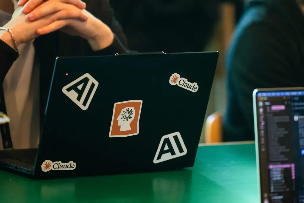 A laptop covered in AI and Claude stickers sits on a desk while someone clasps their hands behind it
