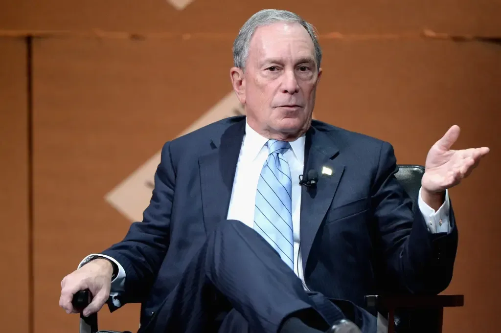 Michael Bloomberg gestures with one raised hand while seated on stage, wearing a dark suit and light blue striped tie