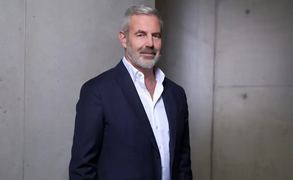 Stefan Klebert, wearing a dark blazer over an open-collar white shirt, stands smiling in front of a concrete wall