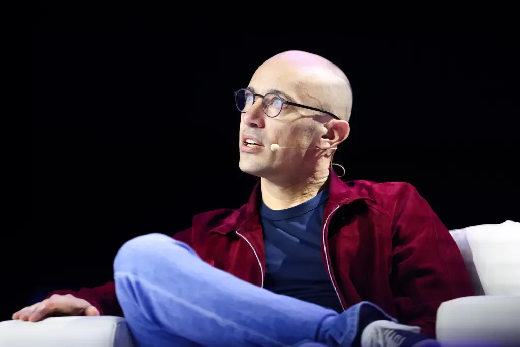 Tobias Lutke sits cross-legged in an armchair on a dark stage, wearing a maroon jacket and a headset microphone, gesturing as he speaks