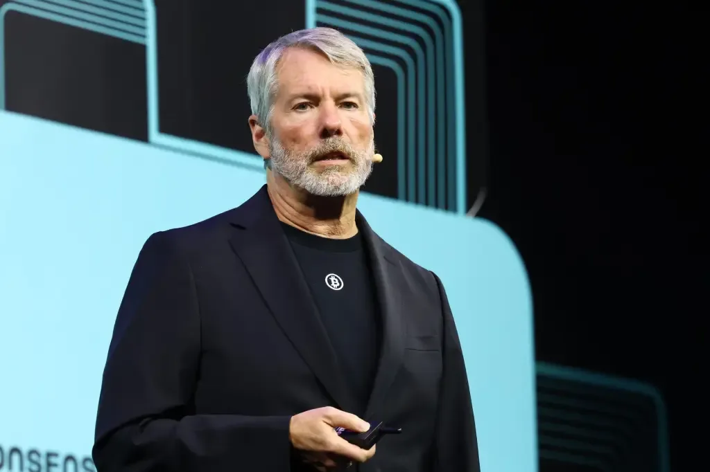 Michael Saylor, wearing a black jacket over a black t-shirt with a Bitcoin logo pin, speaks on stage with a wireless microphone