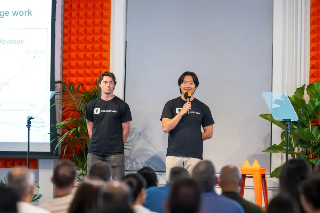 Two Halluminate cofounders, both wearing black Halluminate t-shirts, present to a seated audience with a revenue growth chart displayed on a screen behind them