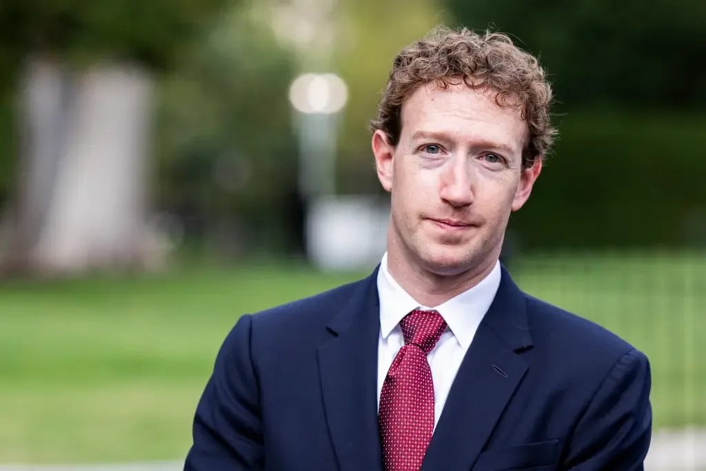 Mark Zuckerberg, wearing a dark suit and red tie, stands outdoors in front of greenery