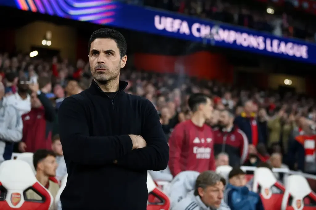 Mikel Arteta stands with arms crossed on the touchline in front of a packed stadium crowd, with a UEFA Champions League sign visible behind him