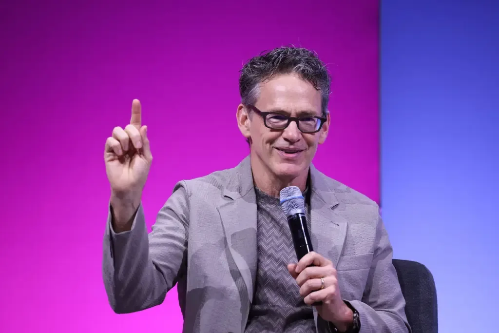 Bill Anderson, wearing glasses and a gray blazer, gestures with one finger raised while speaking into a microphone against a pink and blue stage backdrop