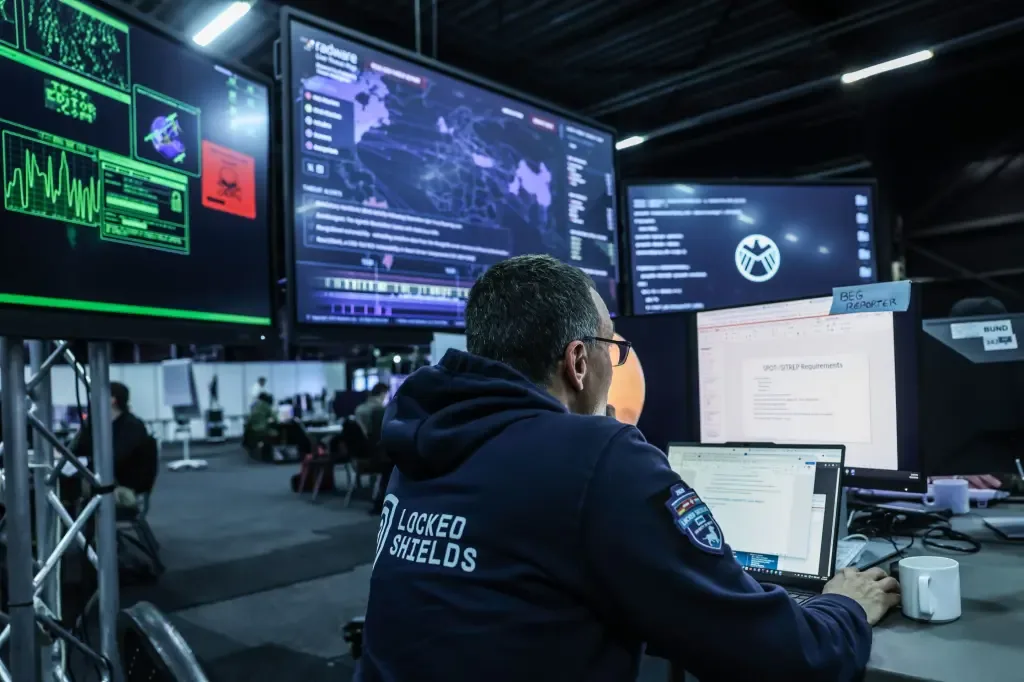A cybersecurity analyst wearing a Locked Shields hoodie monitors multiple large screens displaying threat maps and security data