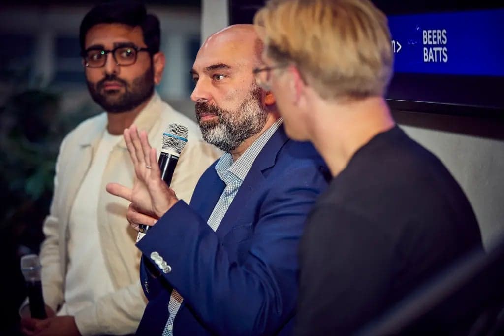 Three men in business attire talk during a panel discussion, one holding a microphone and gesturing, with a screen reading Beers Batts in the background