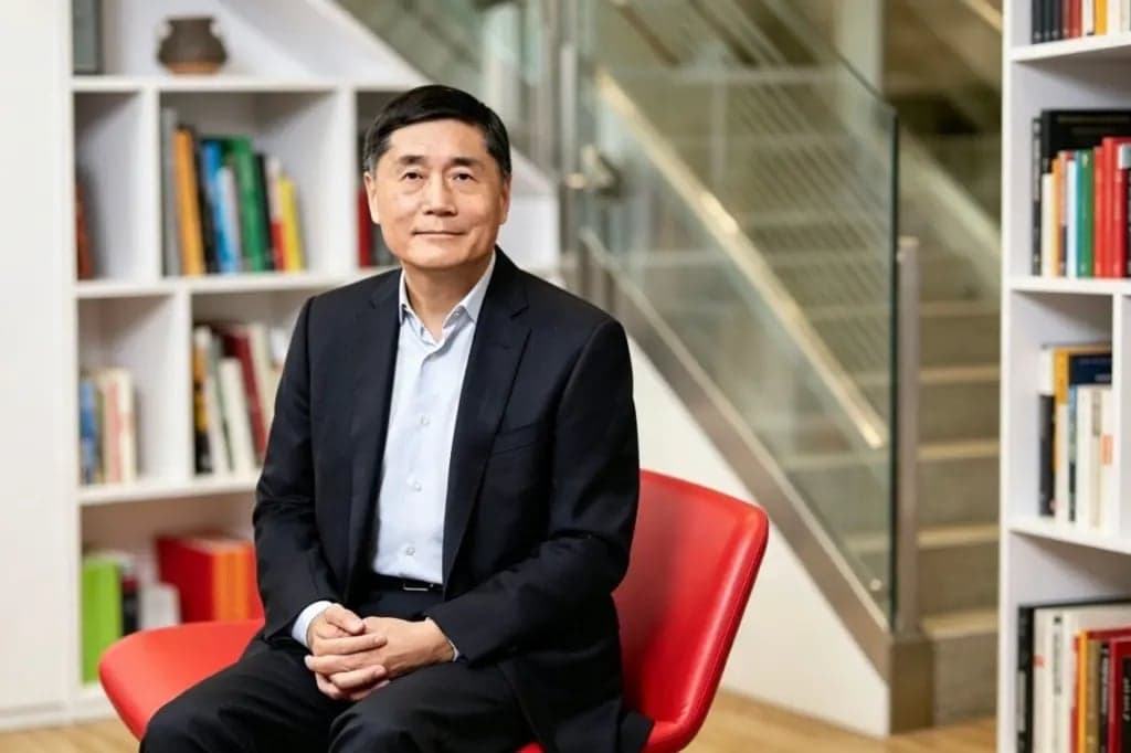 Ming Lu, wearing a dark suit, sits in a red chair in front of bookshelves with a staircase visible behind him