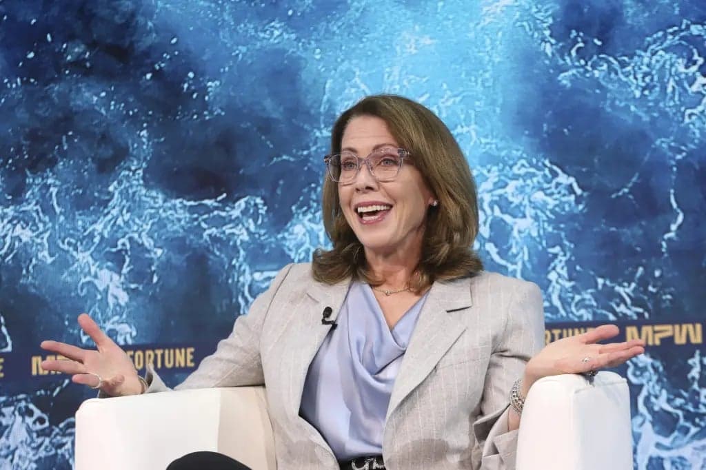A woman wearing glasses and a gray blazer laughs while gesturing with both hands open, seated against a blue Fortune MPW summit backdrop