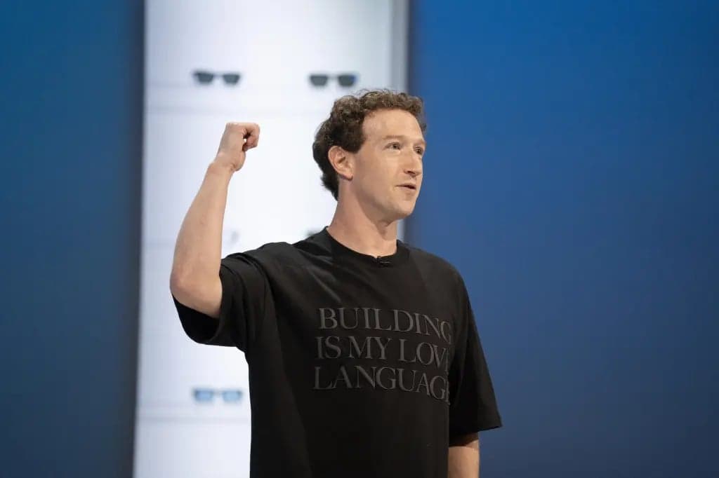 Mark Zuckerberg, wearing a black t-shirt reading Building Is My Love Language, raises his fist while speaking on stage in front of a blue backdrop