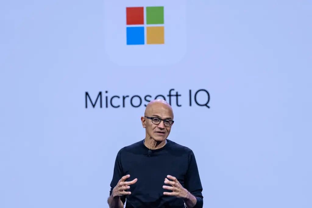 Satya Nadella, wearing a black sweater and glasses, gestures with both hands while speaking in front of a Microsoft IQ sign