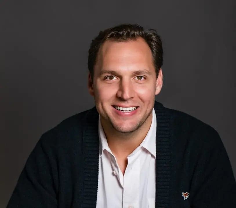 Dane Stevens, wearing a dark cardigan over a white collared shirt, smiles at the camera against a gray background