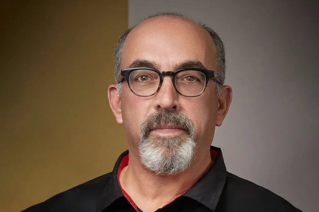 Steven Silverstein, wearing glasses and a black collared shirt, looks at the camera against a gold and gray background