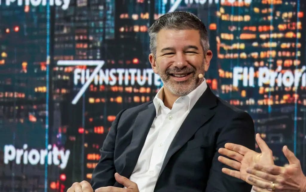 Travis Kalanick, wearing a dark blazer over a white shirt, smiles and gestures with both hands in front of a backdrop of city lights