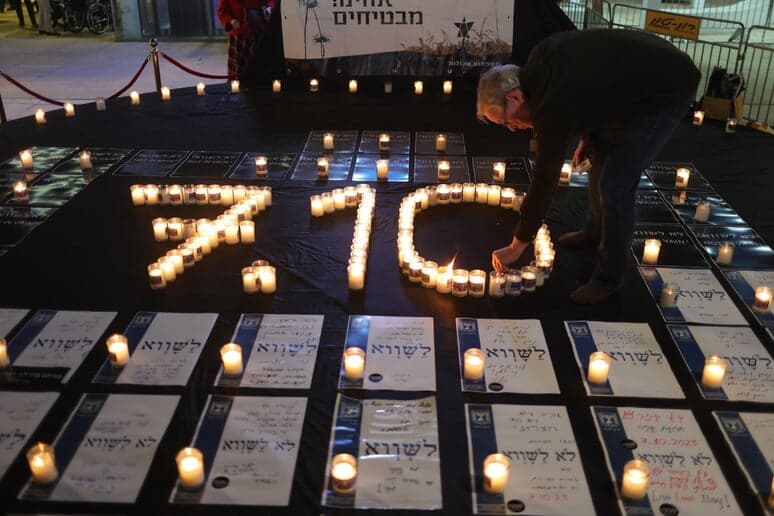 Candles arranged to spell 7.10 surrounded by printed memorial signs in Hebrew, as a man leans over to light one at a nighttime vigil